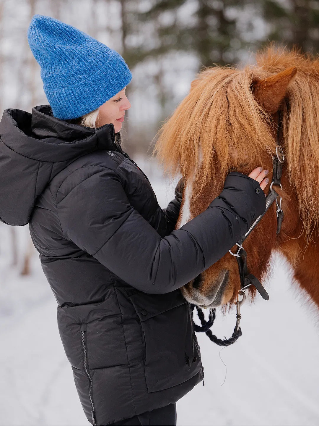 A woman in a Uhip Arctic Puffer Jacket Jet Black and a bright blue knit beanie gently holds a brown horse in a snowy outdoor setting.