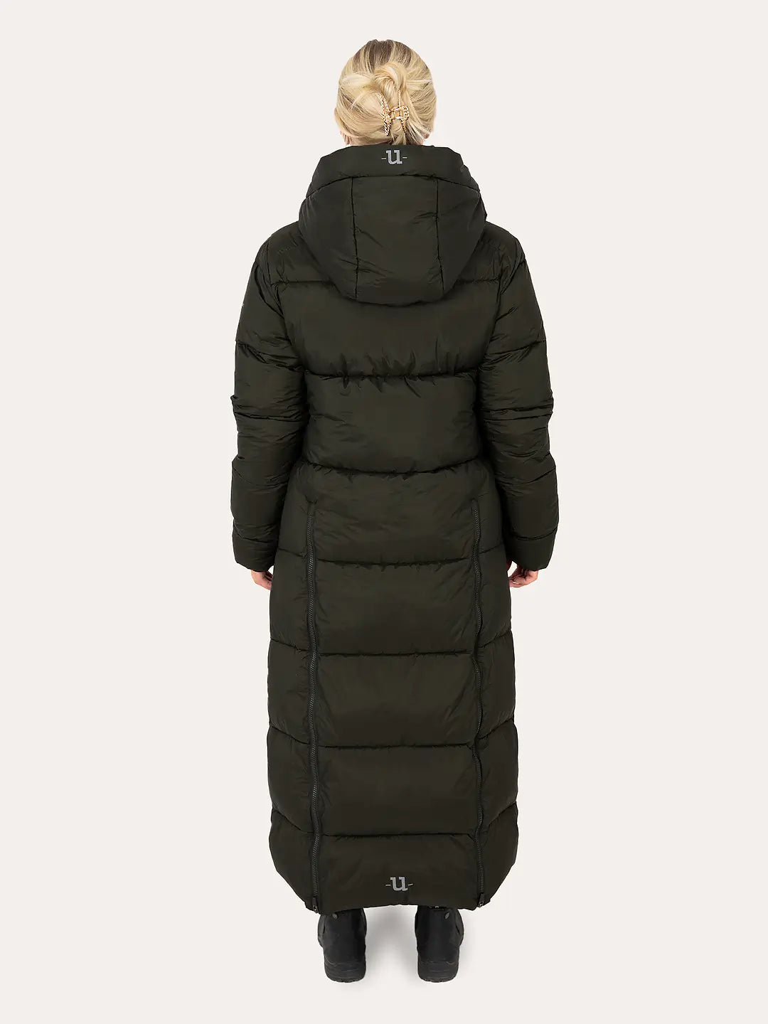 Uhip arctic puffer coat extended dark green. A woman viewed from behind wearing a long, dark olive green puffer coat with a hood, paired with black boots, against a light gray studio background.