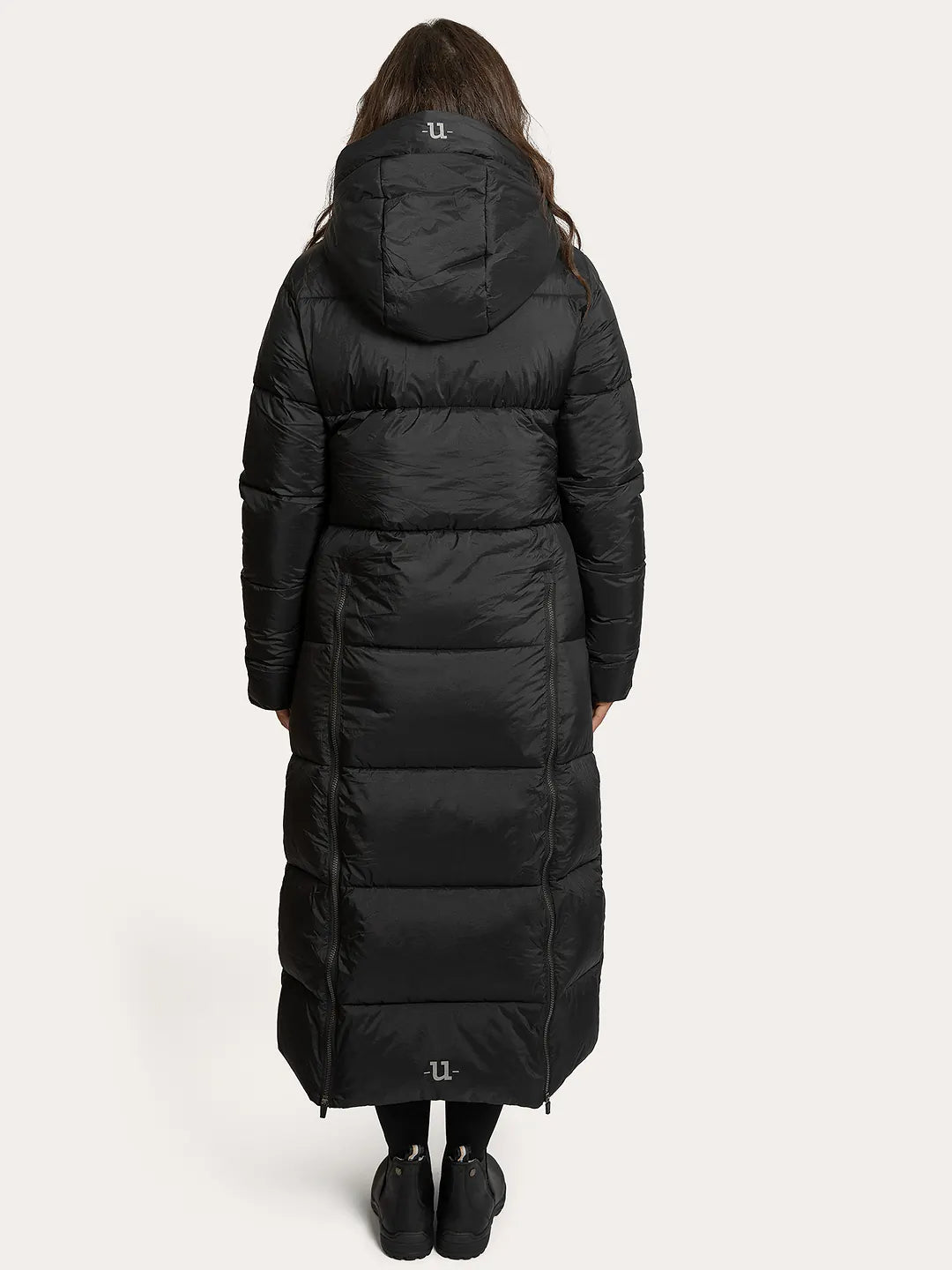 A model viewed from the back wearing a Uhip arctic puffer coat extended Black with horizontal quilting, black leggings, and black boots against a light grey studio background.