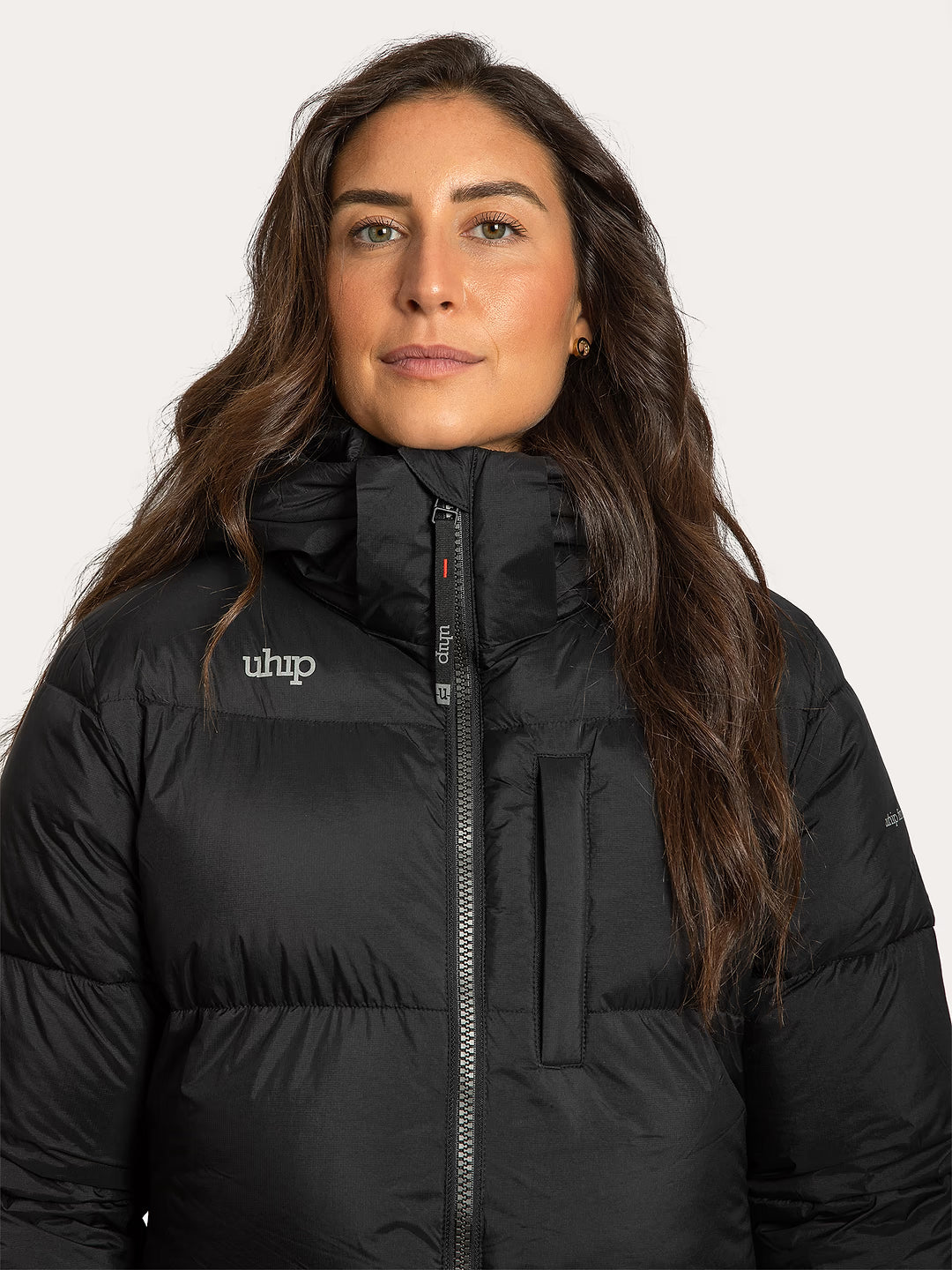 A woman wearing a Uhip arctic puffer coat extended Black with a high collar and front zipper, shown from the chest up against a light grey studio background. The jacket has a visible white reflective 'Uhip' logo on the left chest.