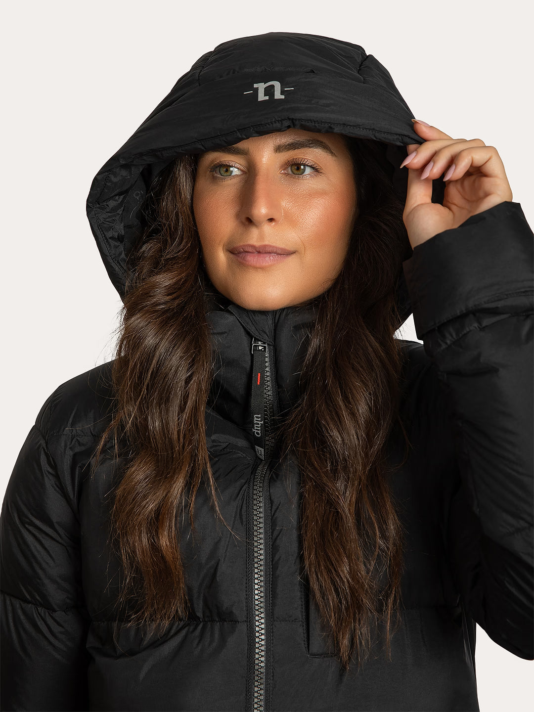 A woman wearing a Uhip arctic puffer coat extended Black and matching detachable black hood, holding up the edge of the hood. The hood features a small white reflective stylized 'U' for Uhip logo.
