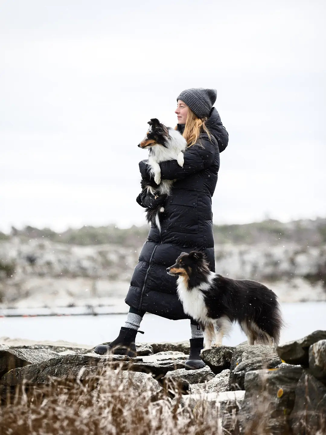 A woman wearing a Uhip arctic puffer coat extended Black grey beanie, and black boots stands on rocks outdoors, holding a small black-and-white dog while another black-and-white dog stands beside her. The background shows a blurred natural landscape with water and sky.