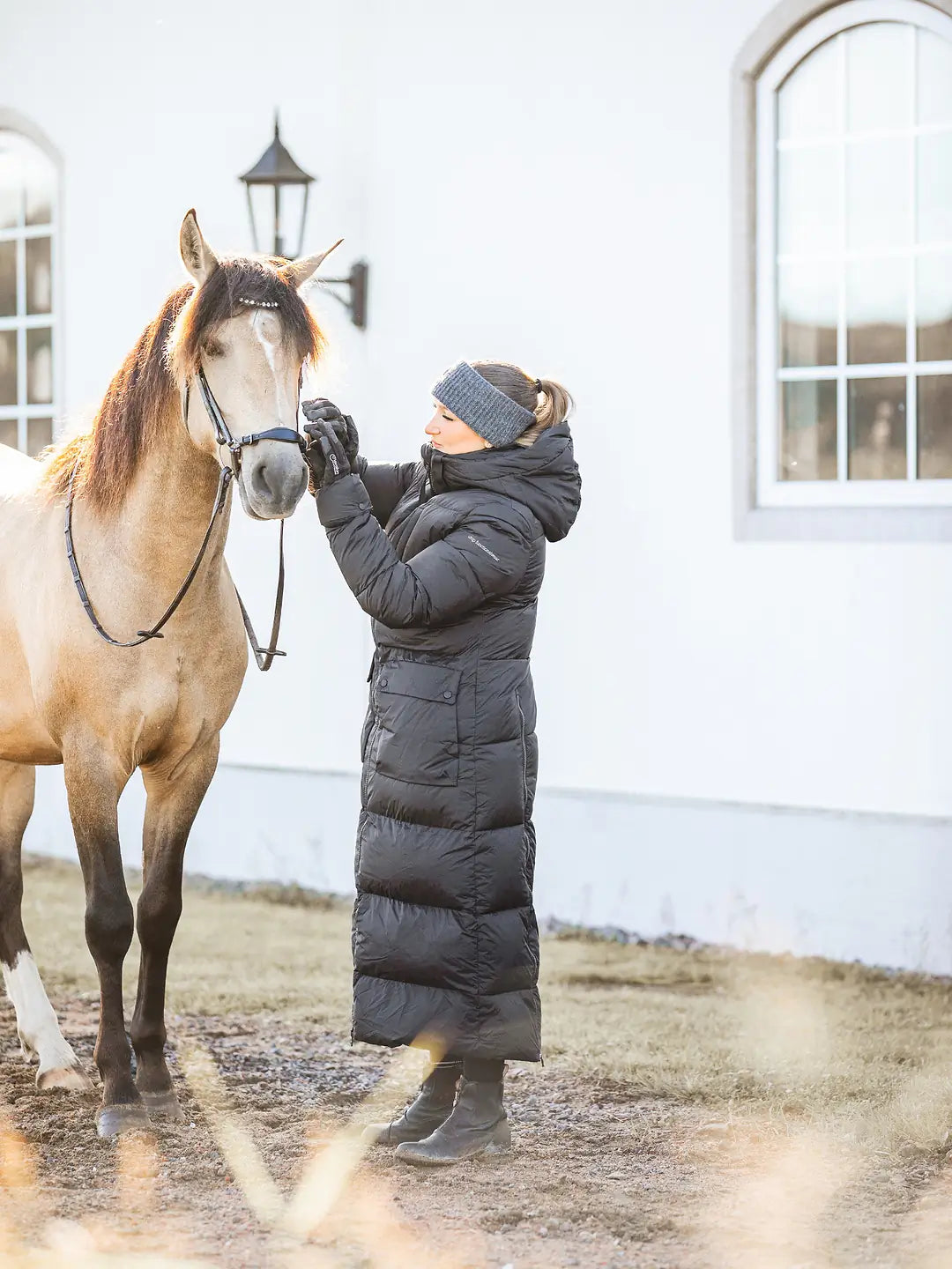 A woman in a Uhip arctic puffer coat extended Black , grey Uhip alpaca beanie, and black gloves is adjusting the bridle on a light brown horse in front of a white building.
