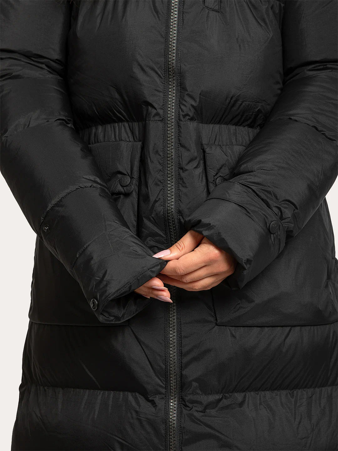 Close-up of a Uhip arctic puffer coat extended Black with horizontal quilting, a front zipper, and a drawstring waist, shown on a model with hands clasped showing extendable sleeves in front.
