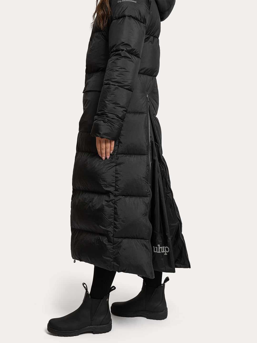 A model wearing a Uhip arctic puffer coat extended Black with a hood and side slit, paired with black ankle boots, shown from a side profile against a light off-white background.