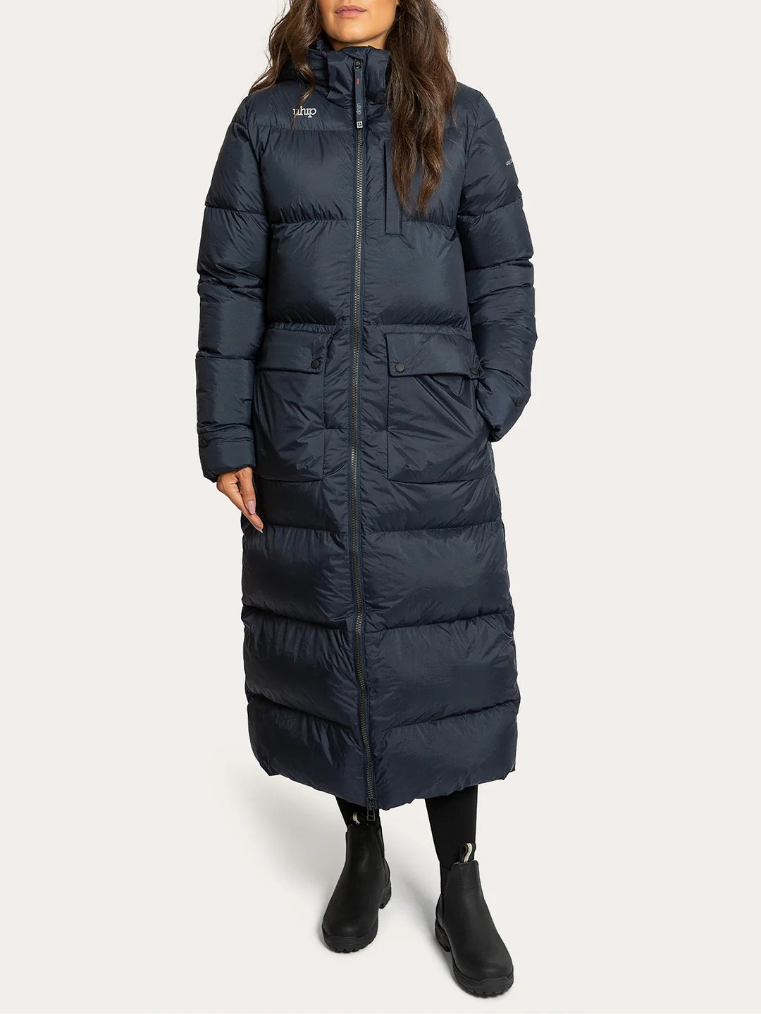 A woman wearing Uhip arctic puffer coat extended eclipse with a high collar, front zipper, and large front pockets, paired with black boots, against a light grey studio background.