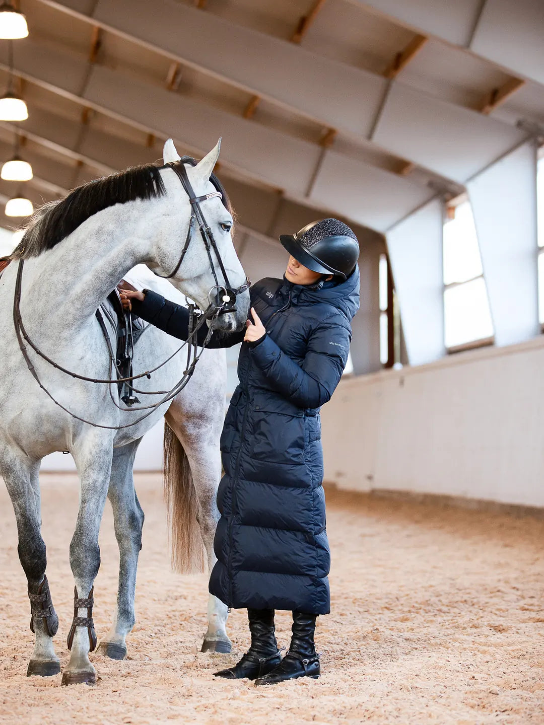 A woman in a Uhip arctic puffer coat extended eclipse and black boots stands in an indoor horse stable next to a dapple grey horse, holding its reins.