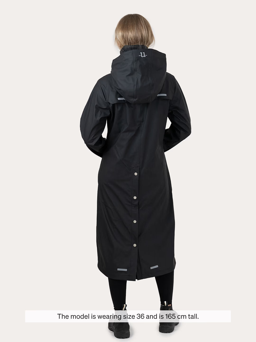 A model shown from the back wearing a Uhip Winnie Raincoat jet black with a buttoned front and reflective details, against a light grey background. A text overlay at the bottom states 'The model is wearing size 36 and is 165 cm tall.