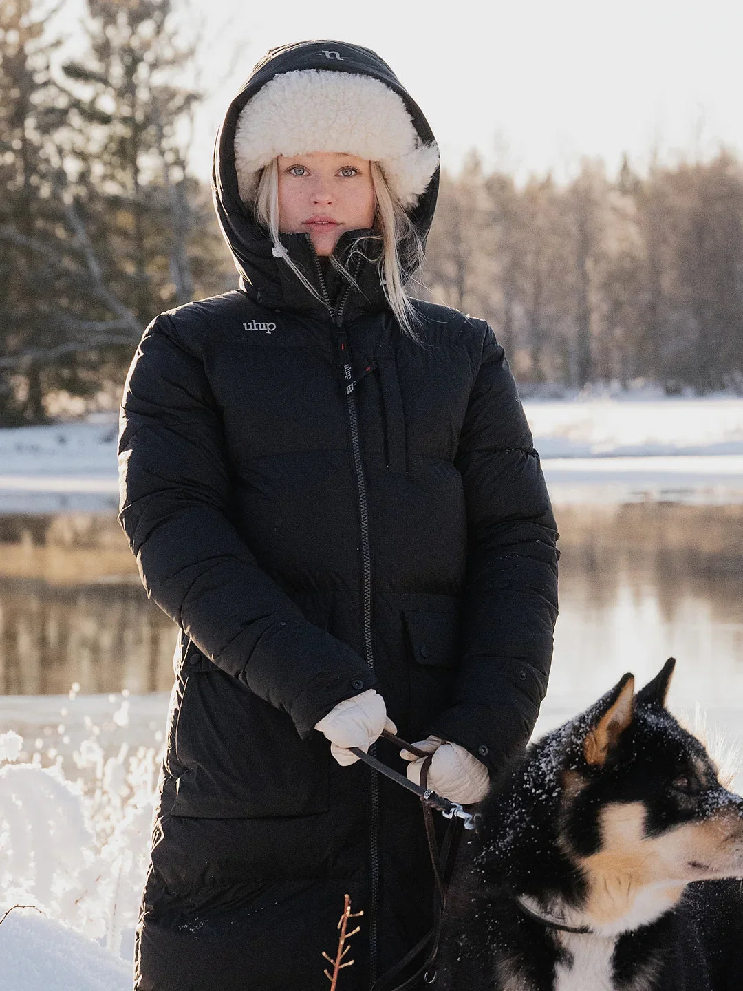 A woman wearing a Uhip arctic puffer coat extended Black with a white faux-fur trimmed hood stands outdoors in the snow next to a black and white dog. The coat has a small white 'uhip' reflective logo on the left chest.
