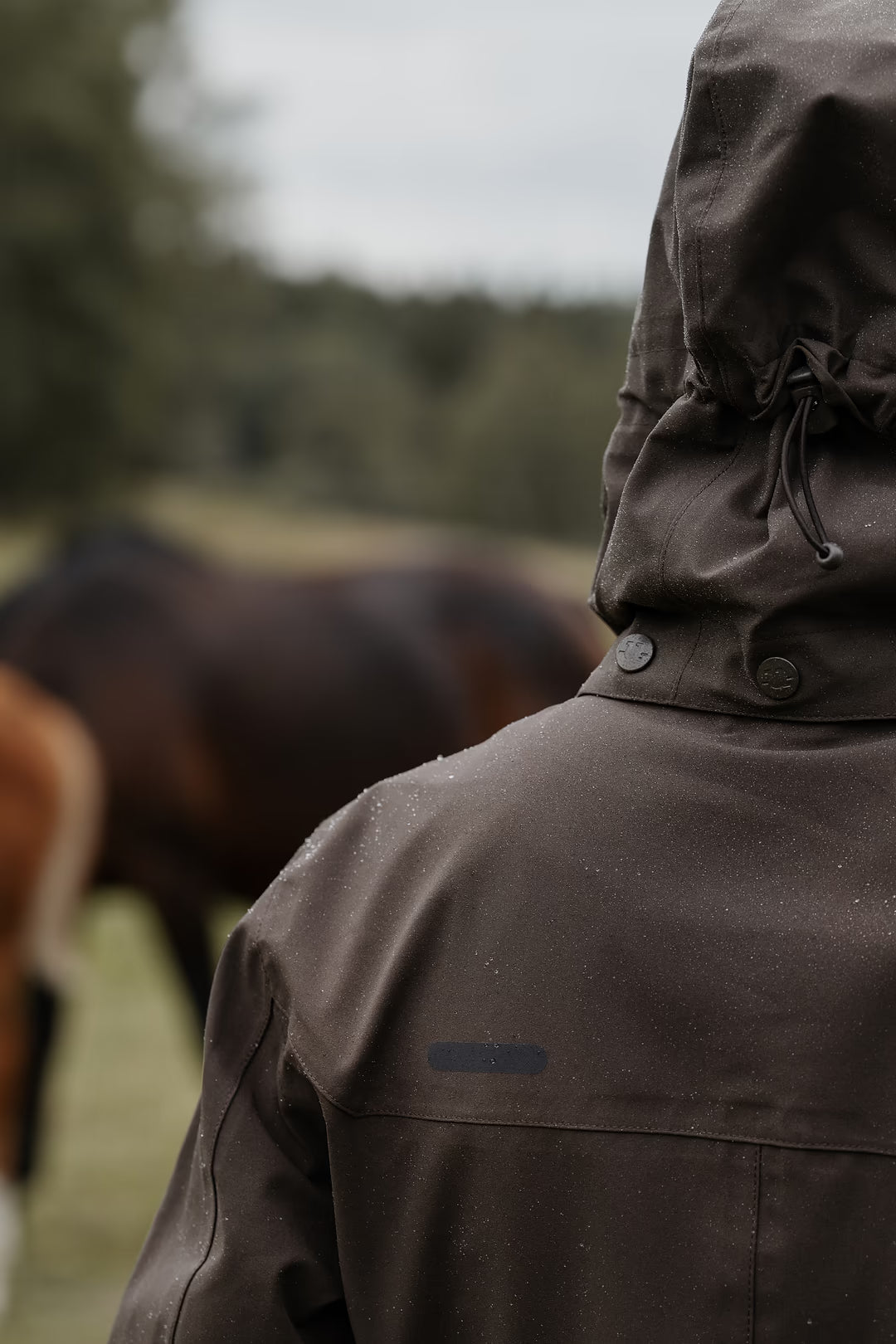 A person viewed from behind wearing a  Uhip Insulated ash brown raincoat outdoors, with a blurred horse and field in the background.