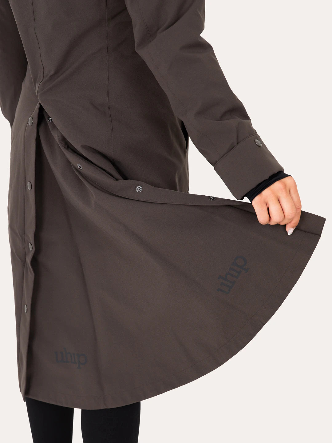 A close-up of a person wearing a Uhip Insulated ash brown raincoat, holding the front flap open to show the interior lining and buttons. The brand name 'uhip' is printed in a subtle dark tone on the lower left of the flap.