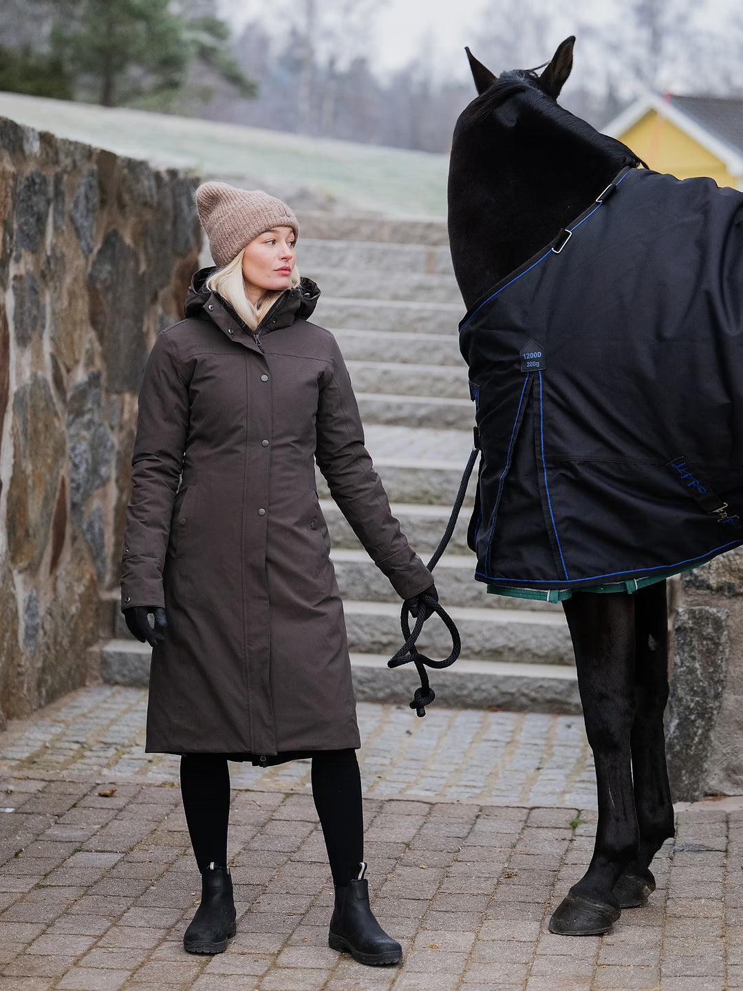 A woman in a Uhip Insulated ash brown raincoat , beige knit Uhip beanie, and black boots stands beside a black horse wearing a black horse blanket with blue trim, on stone steps outdoors.