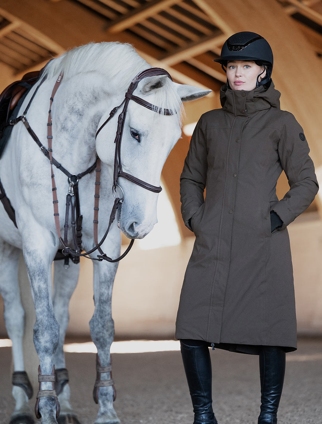 A woman in equestrian attire wearing a Uhip Insulated ash brown raincoat, riding boots, and a black helmet stands next to a white horse in a stable.