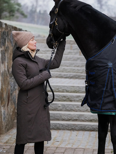 A woman in a Uhip Insulated ash brown raincoat and beige Uhip beanie stands beside a black horse wearing a navy horse blanket with blue piping, set against a stone staircase background.