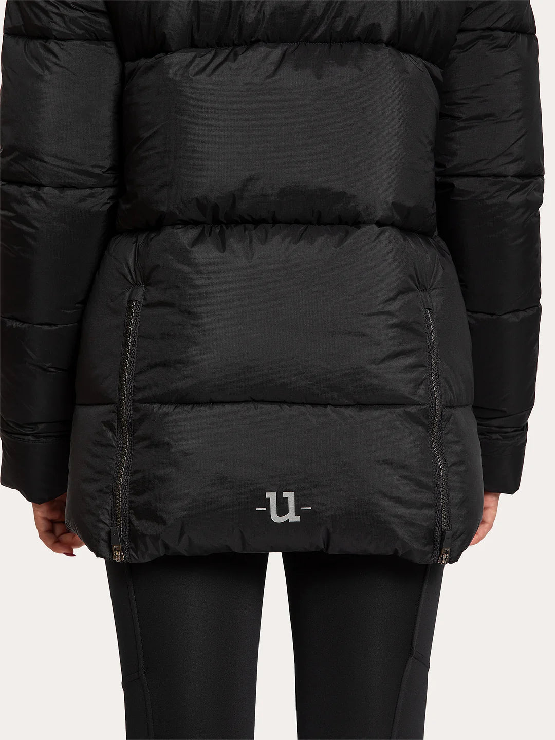 A close-up of a Uhip Arctic Puffer Jacket Jet Black  worn by a model, showing the quilted horizontal stitching, zippered pockets, and a small white logo on the lower front, paired with black leggings against a light grey background.
