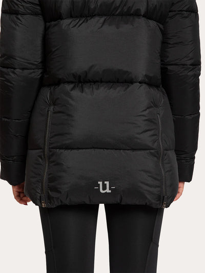 A close-up of a Uhip Arctic Puffer Jacket Jet Black  worn by a model, showing the quilted horizontal stitching, zippered pockets, and a small white logo on the lower front, paired with black leggings against a light grey background.