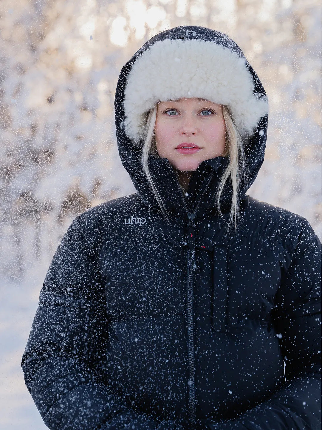 A woman wearing a Uhip Arctic Puffer Jacket Jet Blac with a white faux-fur trimmed hood and a matching black winter hat, standing outdoors in the snow with trees blurred in the background.
