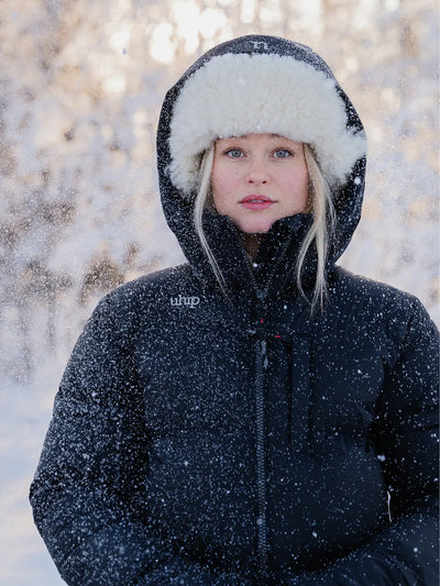 A woman wearing a Uhip Arctic Puffer Jacket Jet Blac with a white faux-fur trimmed hood and a matching black winter hat, standing outdoors in the snow with trees blurred in the background.