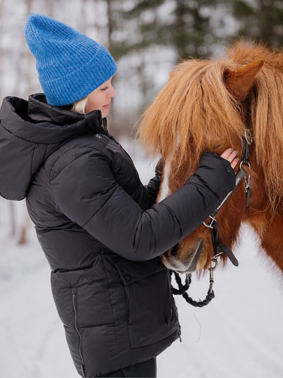 A woman in a Uhip Arctic Puffer Jacket Jet Black and a bright blue knit beanie gently holds a brown horse in a snowy outdoor setting.