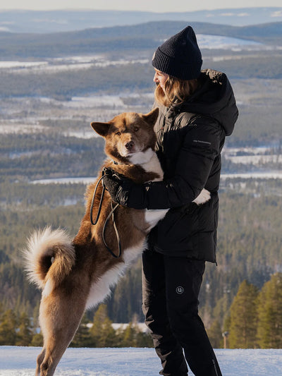 A person in a Uhip Arctic Puffer Jacket Jet Black, beanie, and pants holds a brown and white dog on a snowy mountain with forested hills in the background.