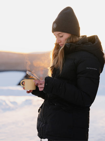 A woman in a Uhip Arctic Puffer Jacket Jet Black and beanie stands outdoors in the snow, holding a steaming mug and looking at a phone. The background shows a softly blurred winter landscape at sunset.