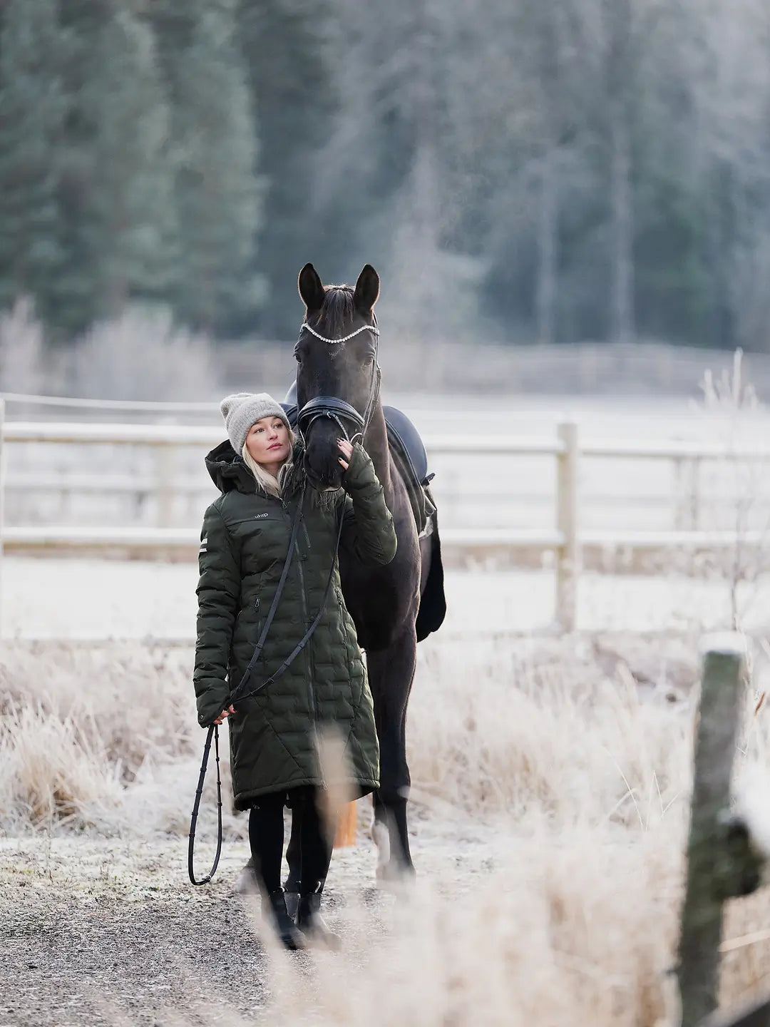 A woman in a Uhip Ice Coat in Dark Green  and grey Uhip beanie walks alongside a dark horse on a frosty path in a rural setting with a wooden fence and trees in the background.