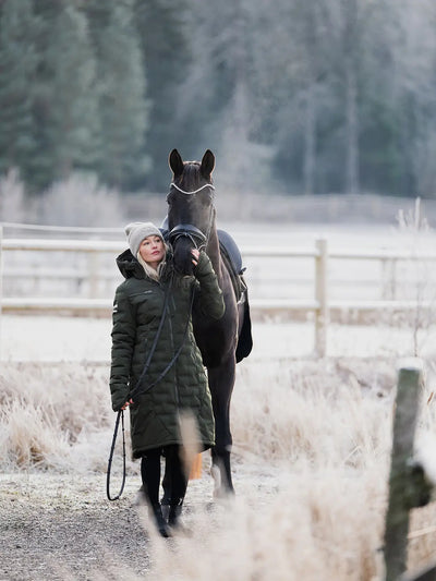 A woman in a Uhip Ice Coat in Dark Green  and grey Uhip beanie walks alongside a dark horse on a frosty path in a rural setting with a wooden fence and trees in the background.