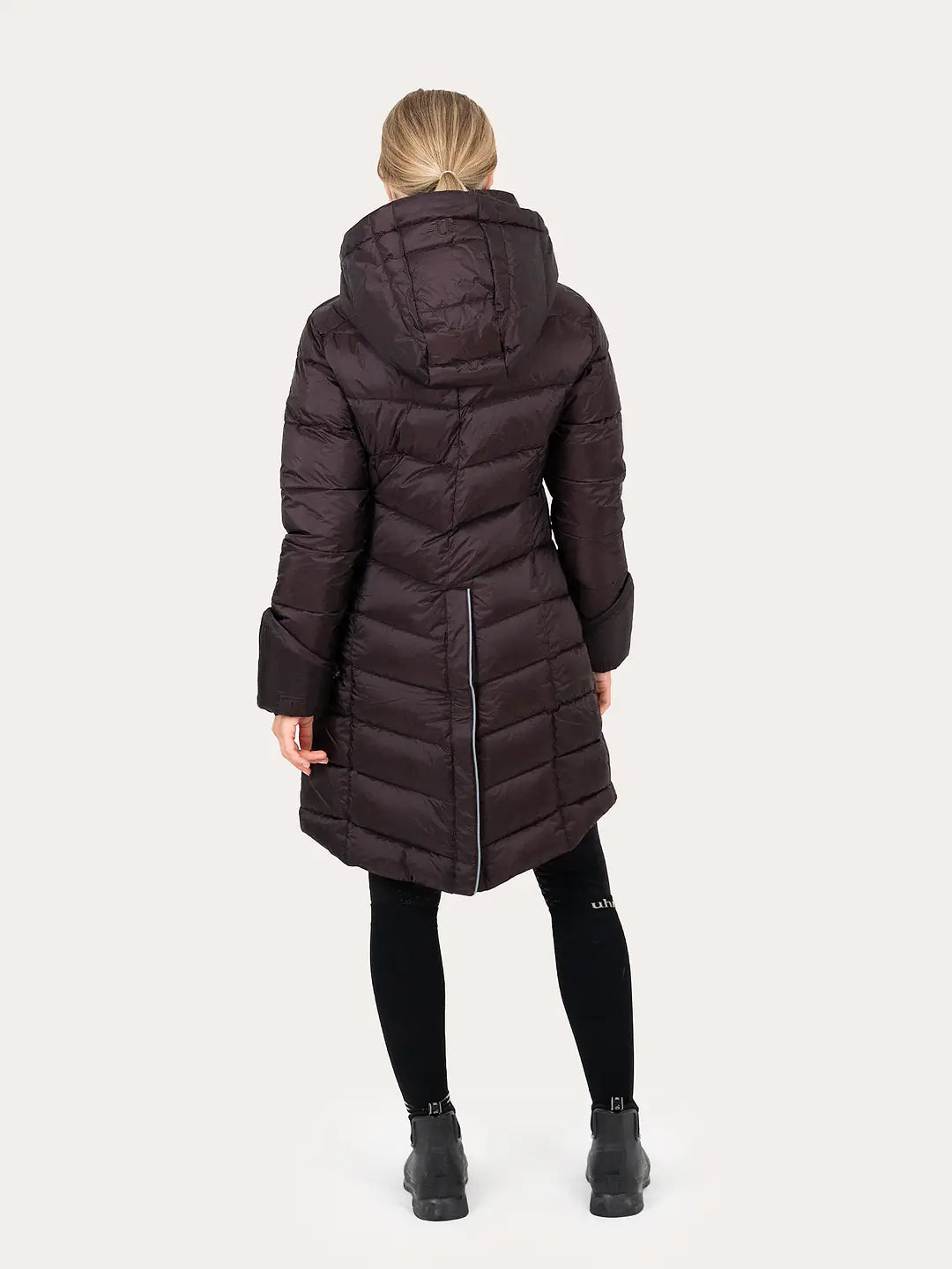 A woman viewed from the back wearing a  Uhip Nordic Mid Coat Chocolate Plum, black leggings, and black boots against a light grey studio background.