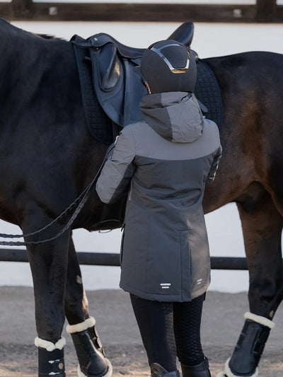A person wearing a Uhip Reflective Jacket 2.0  , black riding tights, and a black riding helmet stands beside a dark horse in an outdoor stable setting.