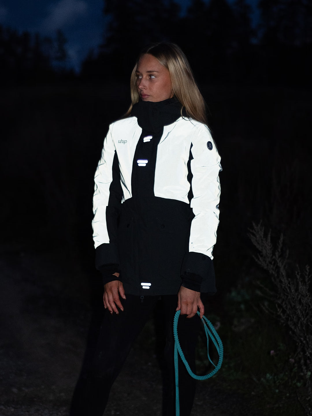 A woman wearing a Uhip Reflective Jacket 2.0  standing outdoors at night holding a teal rope.