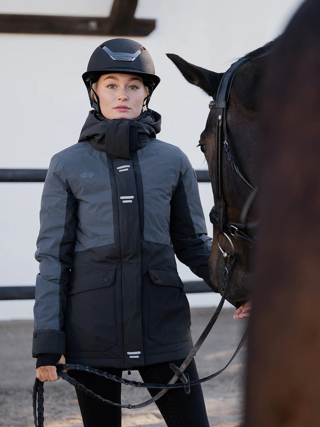 A woman wearing a Uhip Reflective Jacket 2.0  and helmet stands beside a dark horse in an outdoor stable setting.