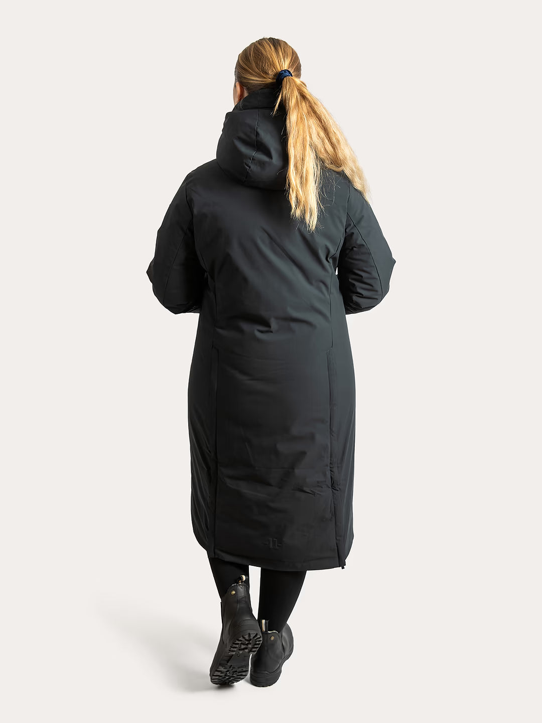 A woman viewed from behind wearing a  Uhip Rime Coat 2.0 Dark Navy, black leggings, and black boots against a plain light grey studio background.