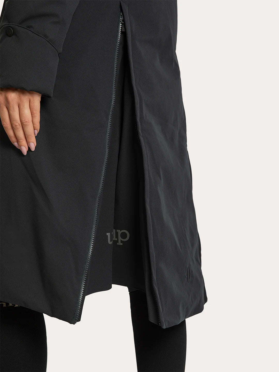 Close-up of a Uhip Rime Coat 2.0 Dark Navy worn by a model, showing a side zipper, sleeve cuff, and a small tonal logo near the hem, against a light off-white background.