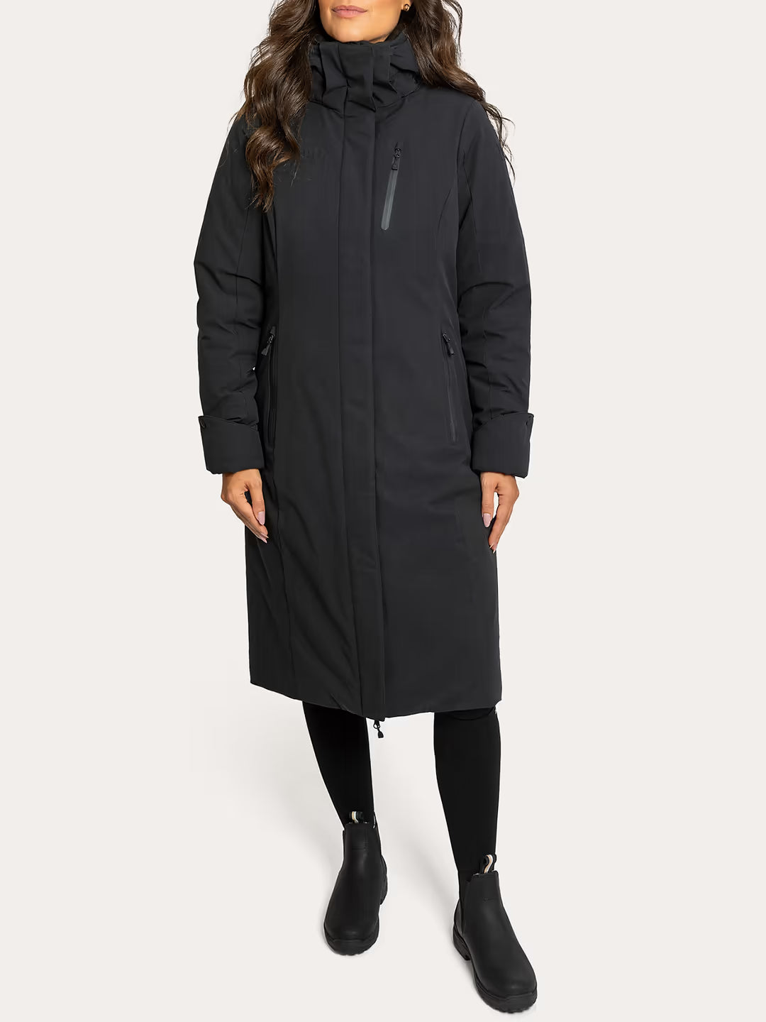 A woman wearing a Uhip Rime Coat 2.0 Dark Navy.t with a high collar, zippered front, and side pockets, paired with black leggings and black ankle boots, against a light grey studio background.