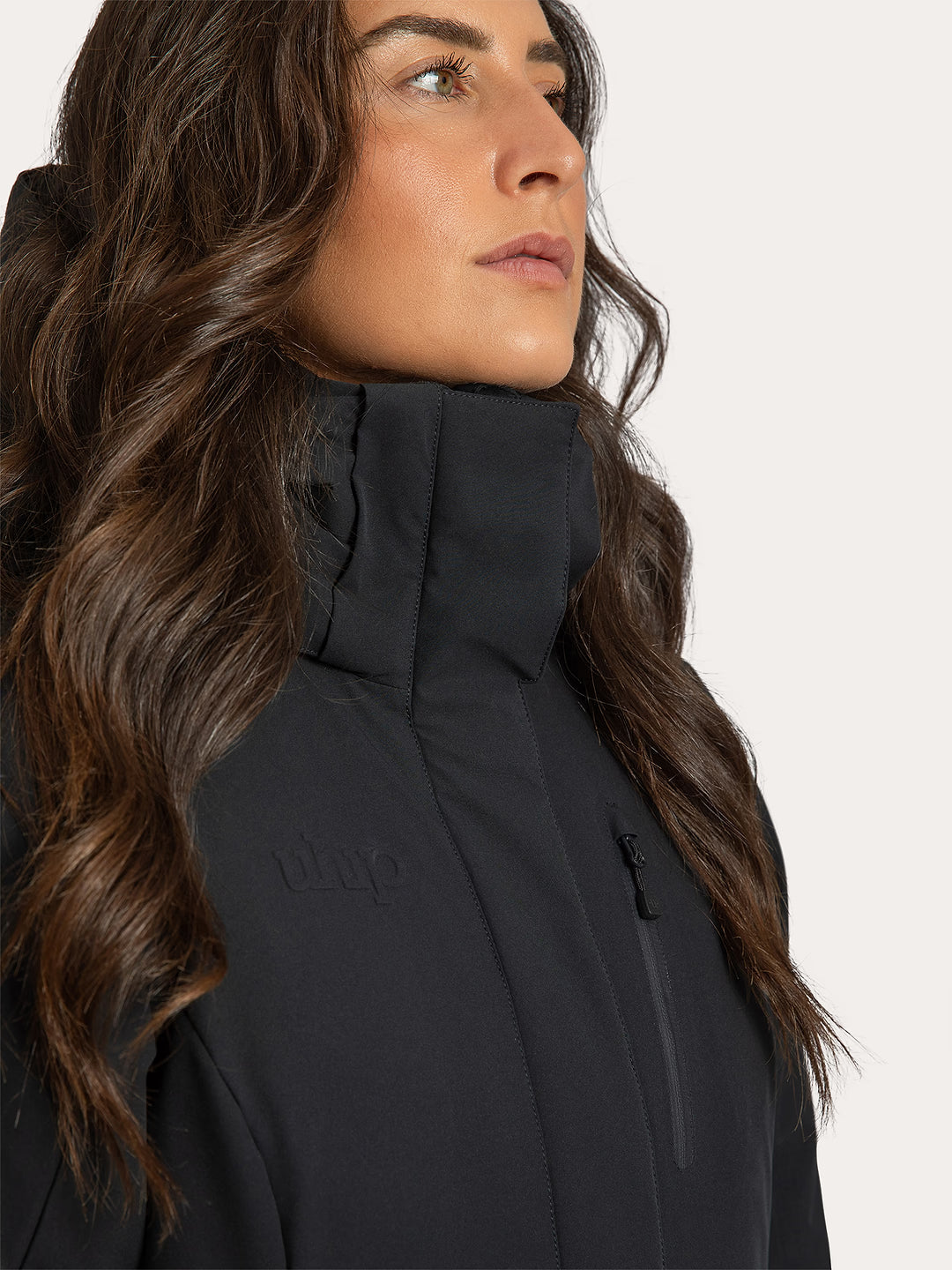 Close-up side profile of a woman wearing a Uhip Rime Coat 2.0 Dark Navy.  with a high collar and subtle Uhip logo on the chest, against a light neutral background.