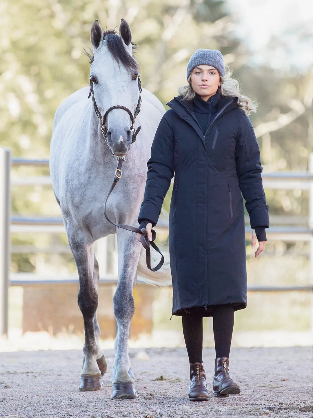 A woman wearing a Uhip Rime Coat 2.0 Dark Navy. Black tights, and a grey Uhip beanie stands beside a dappled grey horse in an outdoor stable or paddock.