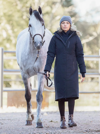 A woman wearing a Uhip Rime Coat 2.0 Dark Navy. Black tights, and a grey Uhip beanie stands beside a dappled grey horse in an outdoor stable or paddock.