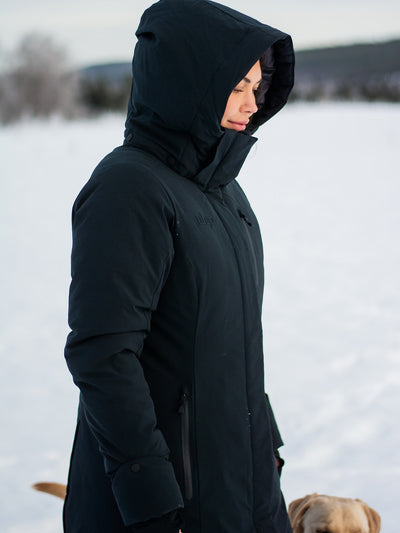 A person wearing a Uhip Rime Coat 2.0 Dark Navy standing outdoors in a snowy landscape, with a dog partially visible at the bottom right.