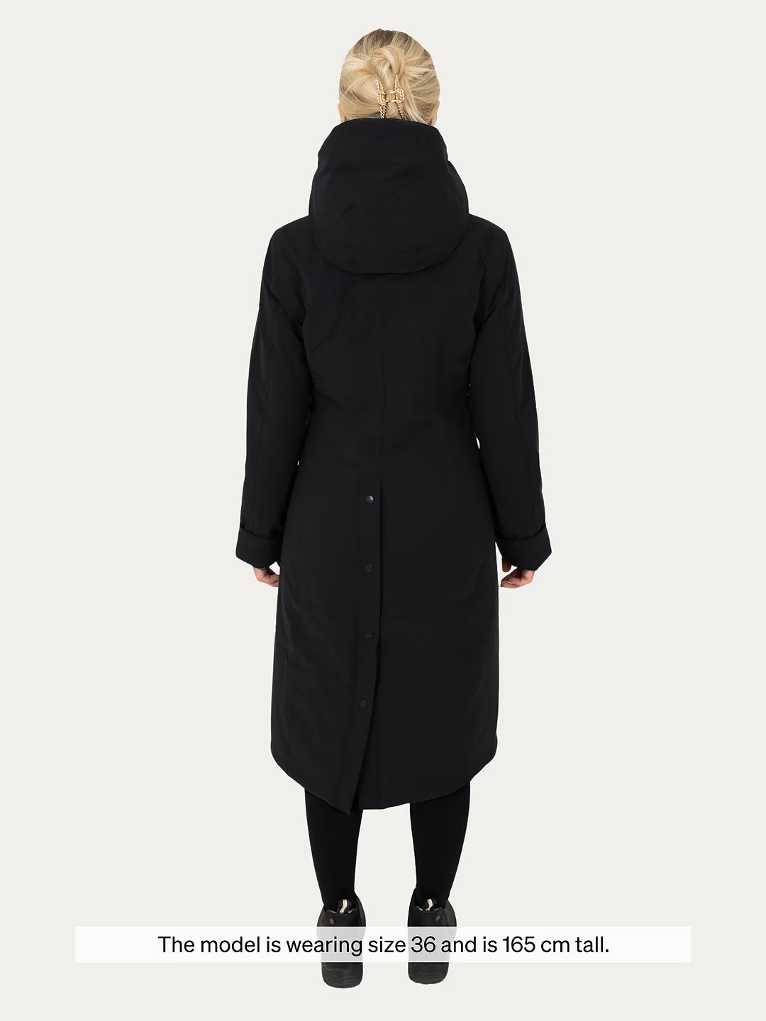 A woman viewed from the back wearing  Uhip Hailey Insulated Jet Black raincoat black leggings, and black shoes against a light gray studio background. A text banner at the bottom reads, 'The model is wearing size 36 and is 165 cm tall.