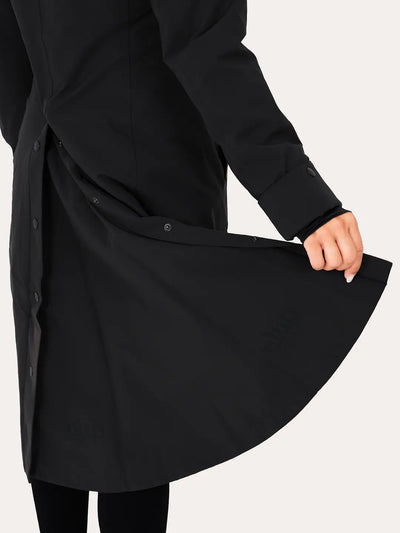 A close-up of a person wearing a  Uhip Hailey Insulated Jet Black raincoat and holding out the side panel to show the fabric and button details, against a plain light background.