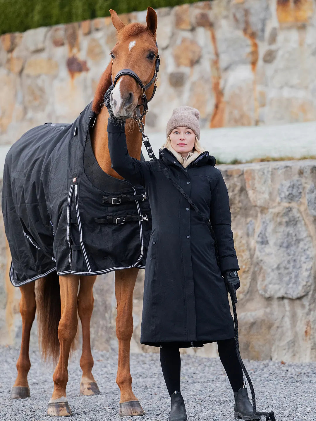 A woman in a Uhip Hailey Insulated Jet Black raincoat  and beige Uhip beanie stands beside a chestnut horse wearing a black horse blanket, with a stone wall and gravel ground in the background.