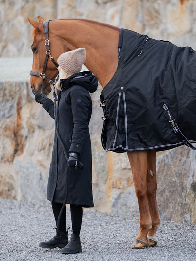 A person in a Uhip Hailey Insulated Jet Black raincoat, black gloves, and a beige beanie stands beside a brown horse wearing a black horse blanket with reflective trim, set against a rocky outdoor background.