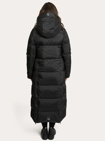 A model viewed from the back wearing a Uhip arctic puffer coat extended Black with horizontal quilting, black leggings, and black boots against a light grey studio background.