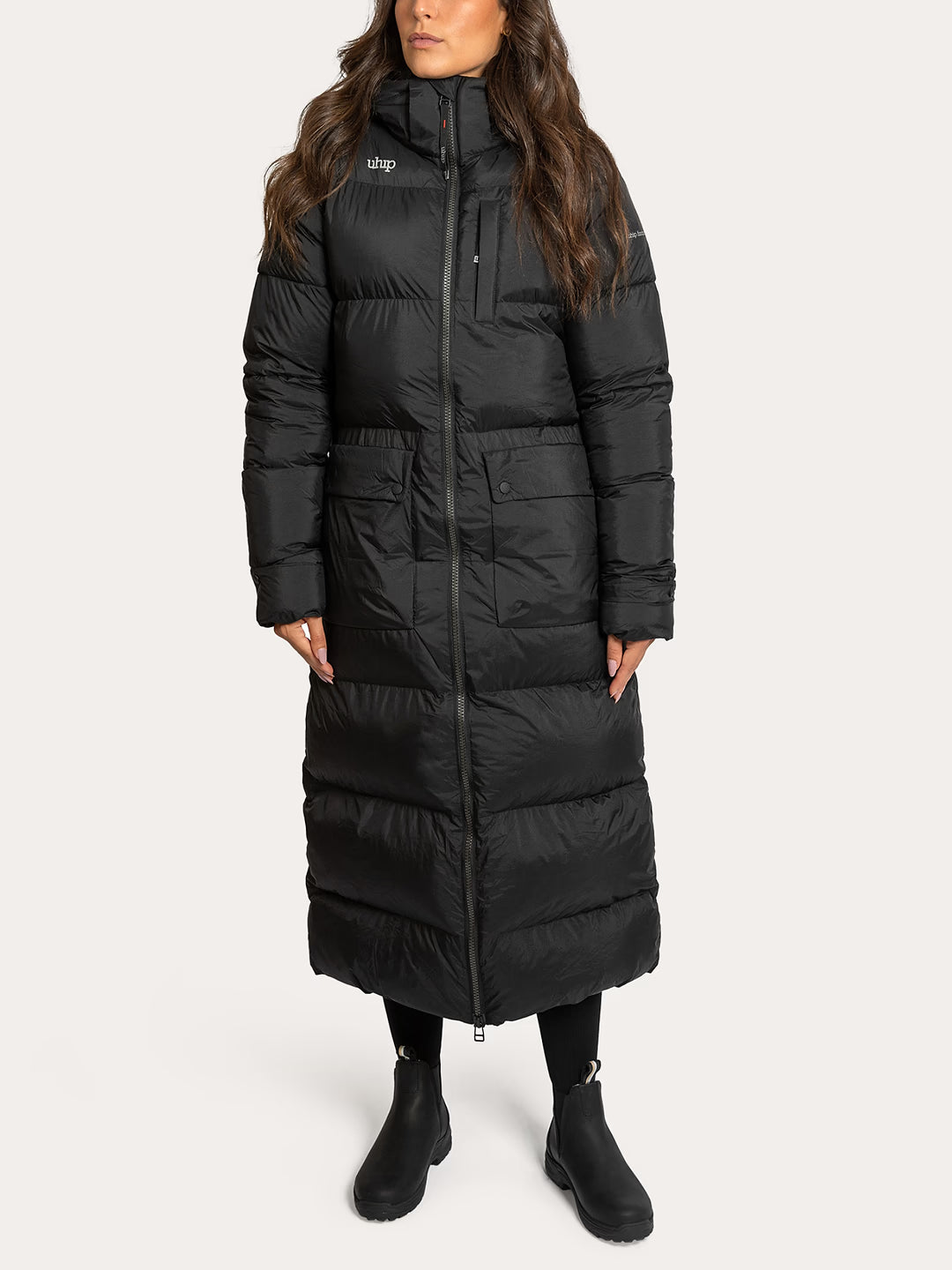 A woman wearing a Uhip arctic puffer coat extended Black  with a high collar, front zipper, and horizontal quilting, paired with black boots, against a light grey studio background.