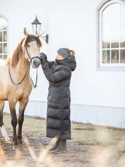 A woman in a Uhip arctic puffer coat extended Black , grey Uhip alpaca beanie, and black gloves is adjusting the bridle on a light brown horse in front of a white building.