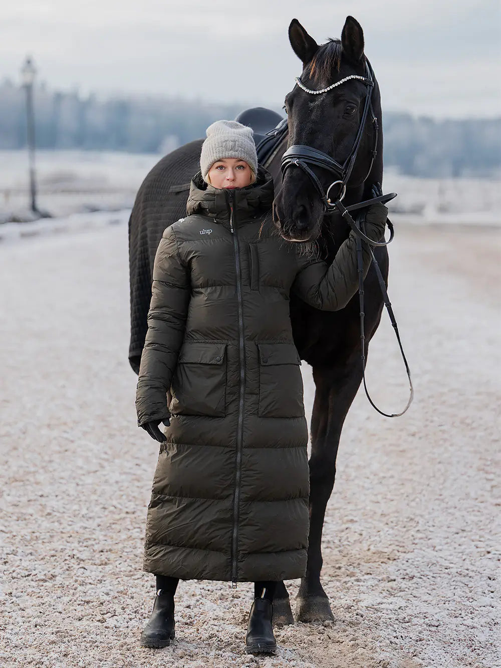 Uhip arctic puffer coat  extended dark green. A woman wearing a long olive green puffer coat and a gray  Uhip beanie stands beside a black horse outdoors on a snowy, open landscape.