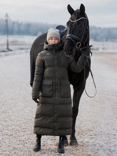 Uhip arctic puffer coat  extended dark green. A woman wearing a long olive green puffer coat and a gray  Uhip beanie stands beside a black horse outdoors on a snowy, open landscape.