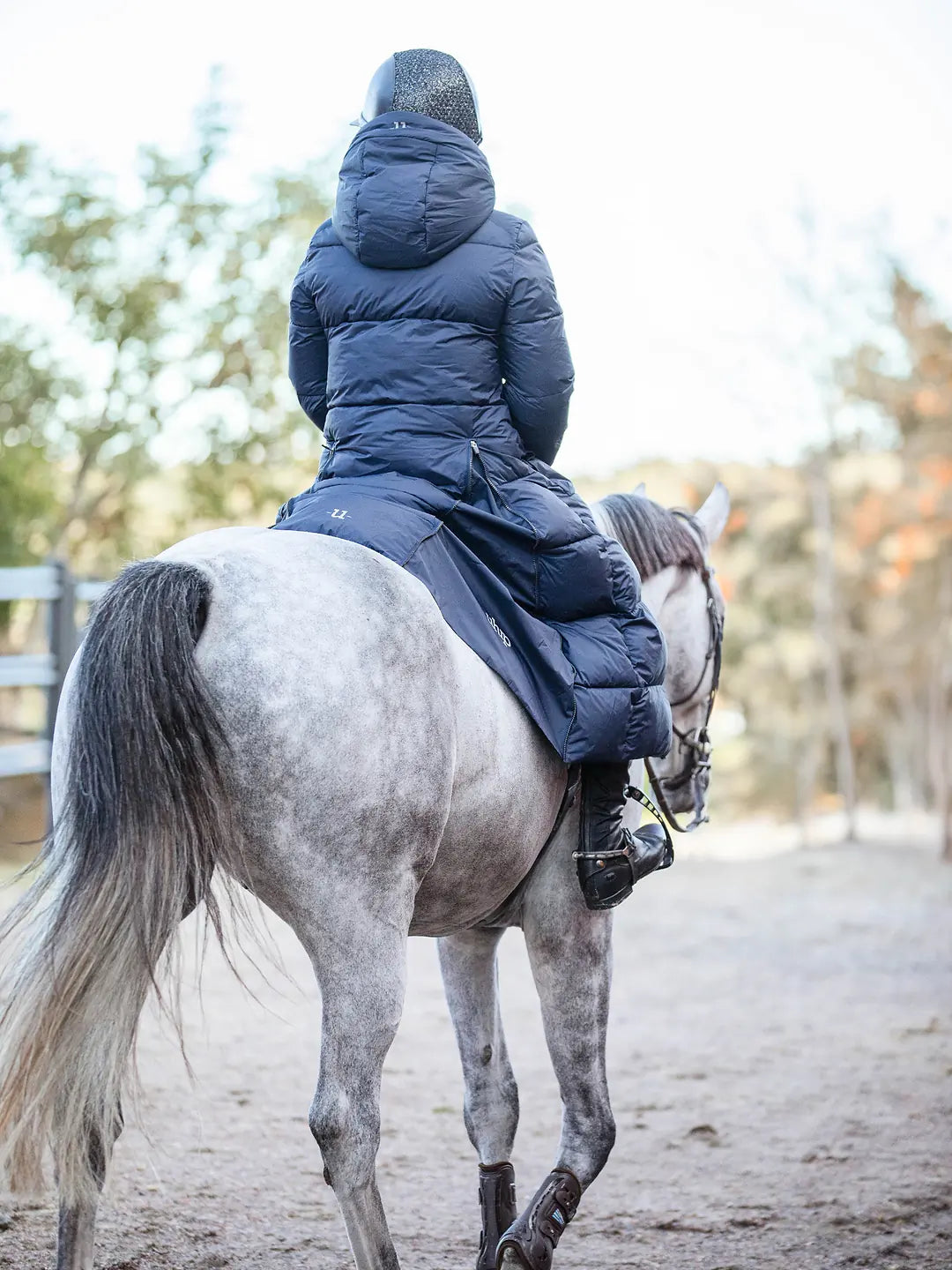 A person wearing a Uhip arctic puffer coat extended  eclipse and riding boots sits on a dapple-grey horse in an outdoor riding arena.