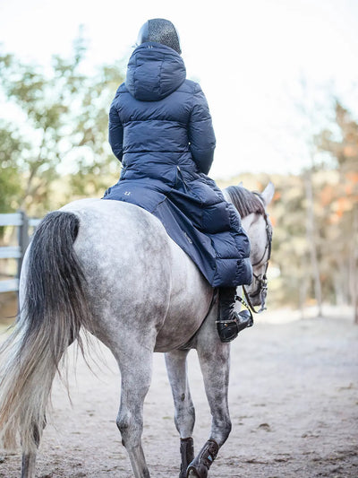 A person wearing a Uhip arctic puffer coat extended  eclipse and riding boots sits on a dapple-grey horse in an outdoor riding arena.