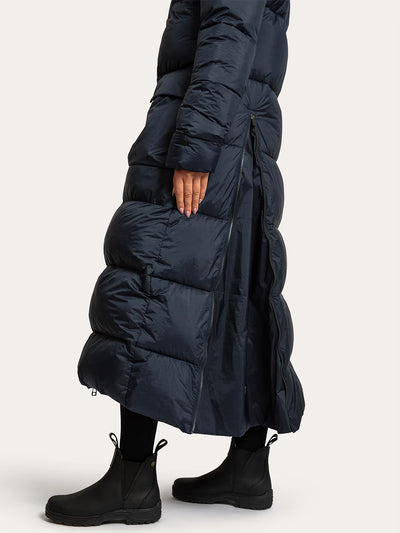 A person wearing a Uhip arctic puffer coat extended  eclipse with horizontal quilting, shown from the side against a light grey studio background. The coat is floor-length and has a side zipper detail, paired with black boots and black leggings.